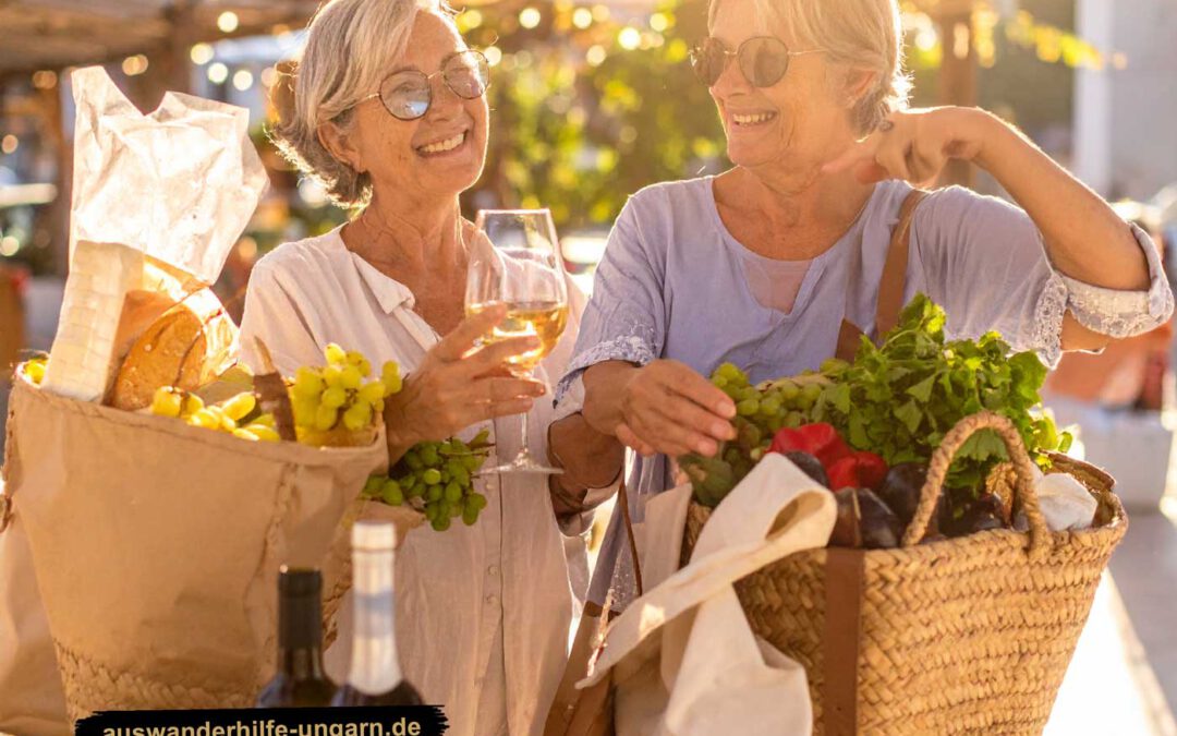 Zwei fröhliche ältere Frauen aus Siklos mit Einkaufskörben voller Gemüse, Brot und Weintrauben lachen auf einem Markt in Ungarn, eine hält ein Glas Wein – Symbol für Lebensfreude und Gemeinschaft.